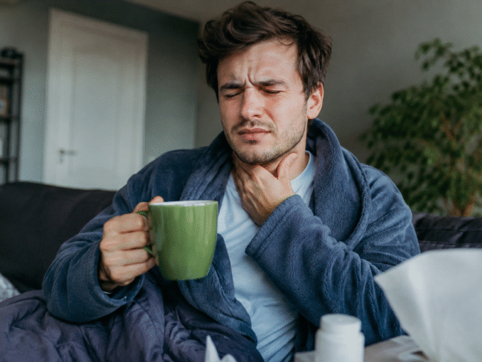 Mann mit Halsschmerzen hält sich den Hals und trinkt eine Tasse Tee