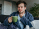 Mann mit Halsschmerzen hält sich den Hals und trinkt eine Tasse Tee