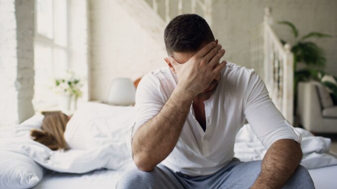 Verzweifelter Mann sitzt im sonnigen Schlafzimmer auf dem Bett und hält sich die Hand vors Gesicht.